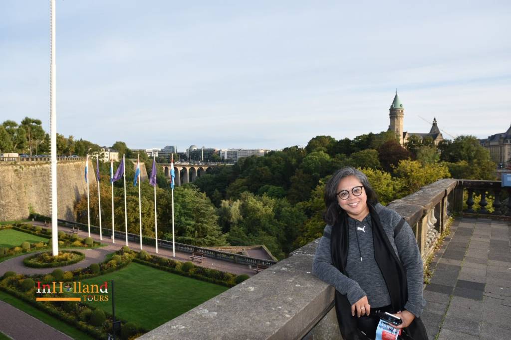 Elly Afriani di Place de la Constitution Luksemburg dengan latar belakang bendera dan Jembatan Adolphe.
