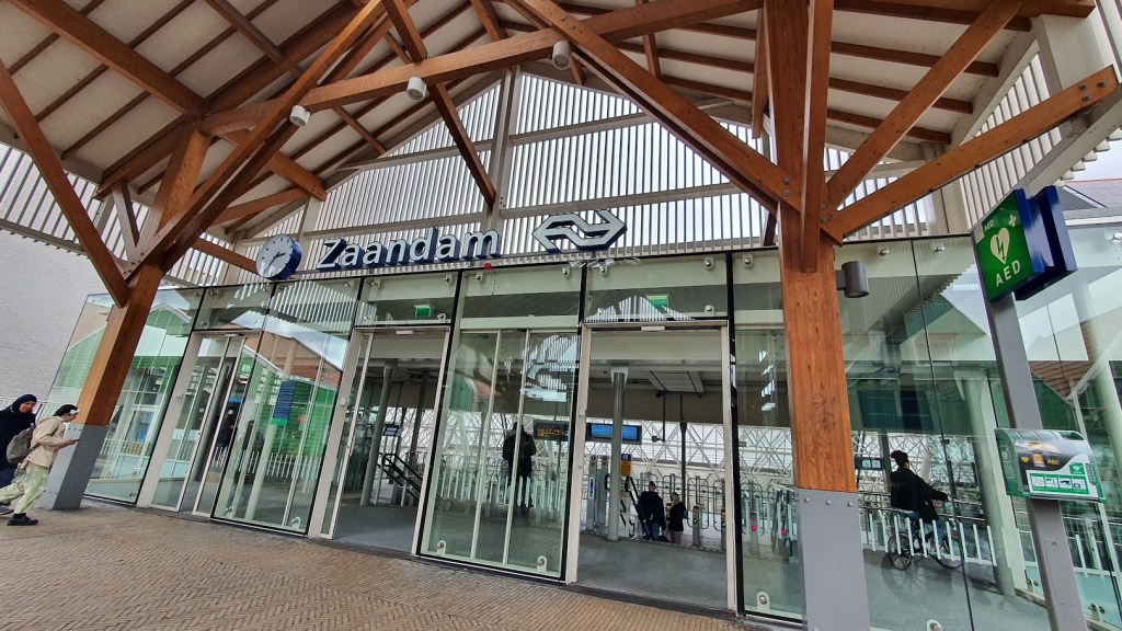 Pintu keluar Stasiun Kereta Api Zaandam dengan latar belakang gedung ikonik bergaya arsitektur rumah kayu tradisional Belanda.